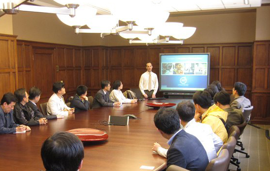 A group of men at a conference table, listening to a man giving a presentation